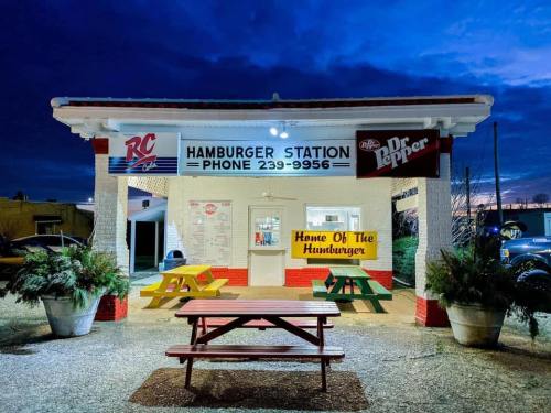 A brightly lit hamburger stand at dusk, featuring colorful picnic tables and signs for RC and Dr Pepper.