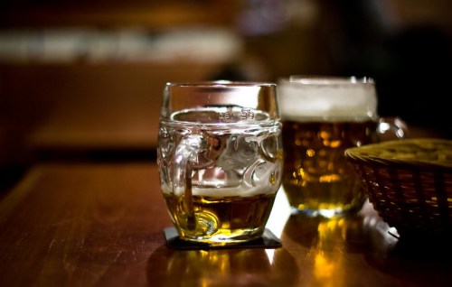 Two glasses of beer on a wooden table, one with foam, next to a basket of snacks. Warm, inviting atmosphere.