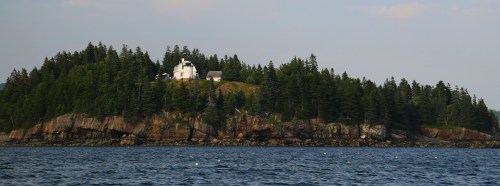A small island with a white house and trees, surrounded by calm water under a clear sky.