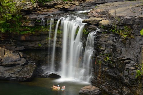 A serene waterfall cascades over rocky cliffs into a pool, with people enjoying the water below.