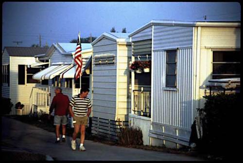 Two men walk past a row of mobile homes, with an American flag displayed on one of the units.