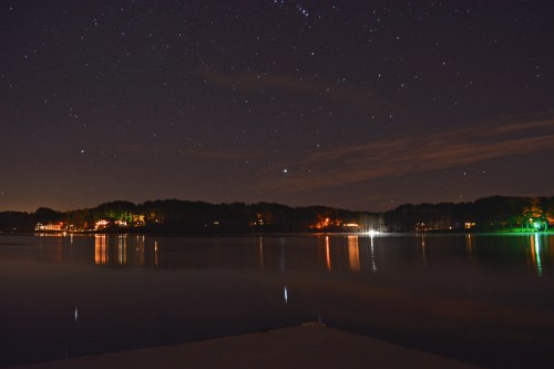 A serene night scene over a calm lake, reflecting stars and distant lights from nearby homes.
