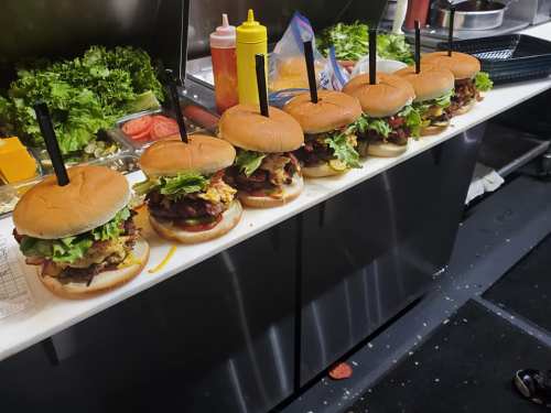 A row of freshly made burgers with lettuce, bacon, and condiments on a countertop, surrounded by fresh ingredients.
