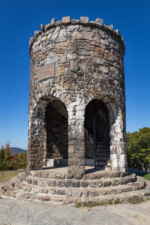 Stone observation tower with arched openings, set against a clear blue sky and surrounded by trees.