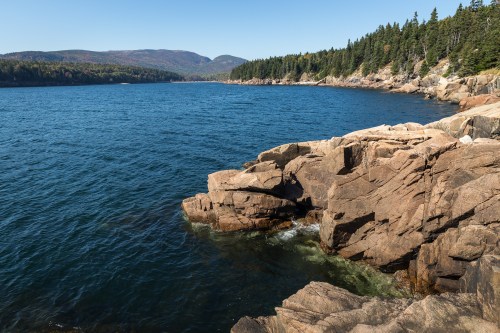 A serene coastal view featuring rocky shores, clear blue water, and lush green forests under a clear sky.
