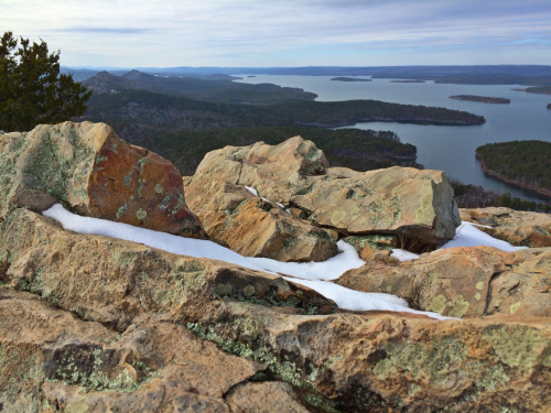 Rocky landscape with patches of snow, overlooking a lake and distant hills under a cloudy sky.
