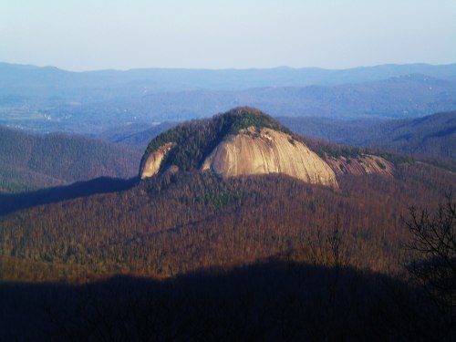 A large, rocky mountain rises above a forested landscape, with rolling hills in the background under a clear sky.