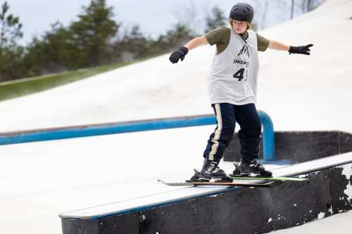 A young skier balances on a rail at a snowy slope, wearing a helmet and protective gear.