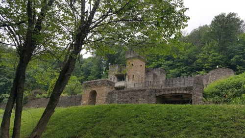 A stone castle surrounded by greenery, with a tower and walls, set against a backdrop of lush trees.