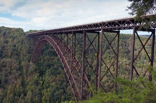 A large, arched metal bridge spans a lush green valley, surrounded by trees under a cloudy sky.