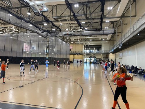 A spacious indoor gym with players practicing softball, some throwing and others catching, in a well-lit environment.