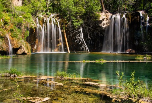 A serene landscape featuring cascading waterfalls flowing into a clear, greenish-blue pool surrounded by lush greenery.