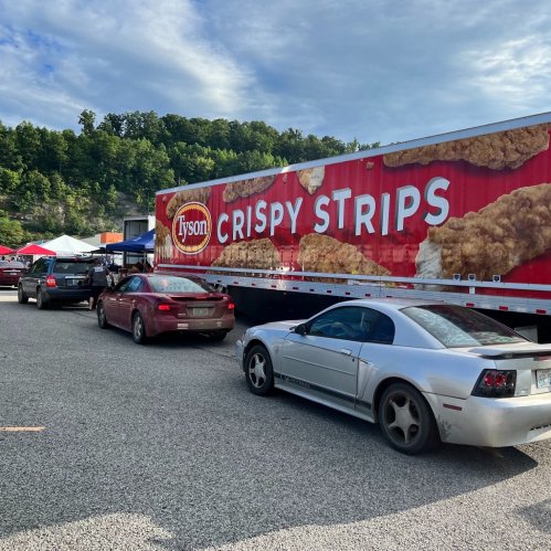 A line of cars waiting near a large Tyson Crispy Strips truck in a gravel lot surrounded by trees.