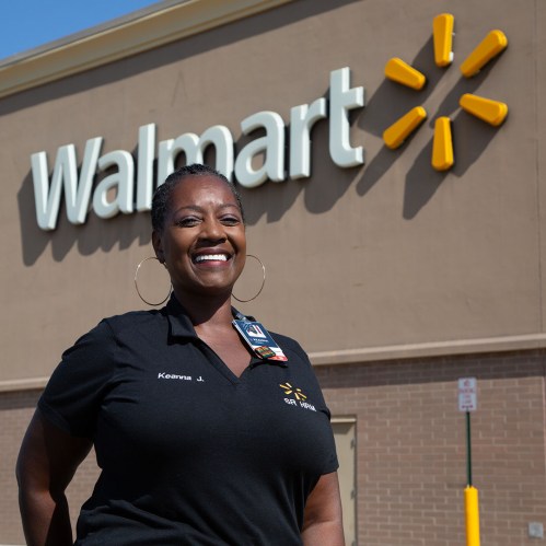 A smiling employee stands in front of a Walmart store, wearing a black shirt with a name tag.