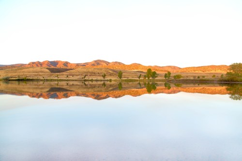 A serene lake reflecting mountains and trees under a clear sky at sunset.