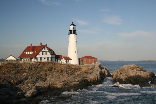 A white lighthouse with a black top stands on rocky shore, beside a red-roofed house, overlooking the ocean.