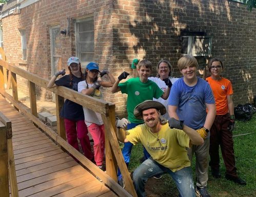 A group of children and adults pose on a wooden ramp, smiling and showing thumbs up, with a brick building in the background.