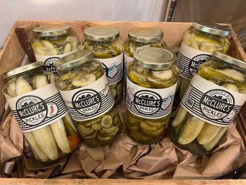 A wooden crate filled with jars of McClure's pickles, featuring various types of pickled cucumbers and peppers.