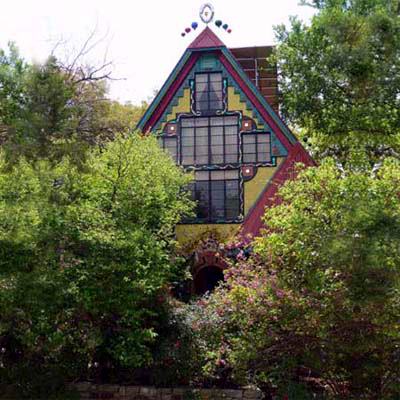 Colorful, whimsical house with a triangular roof, surrounded by lush greenery and vibrant flowers.