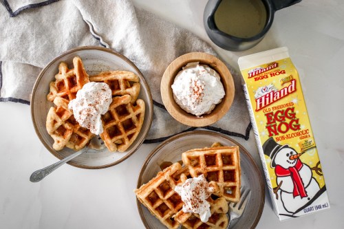 Two plates of waffles topped with whipped cream and nutmeg, alongside a bowl of ice cream and a carton of egg nog.
