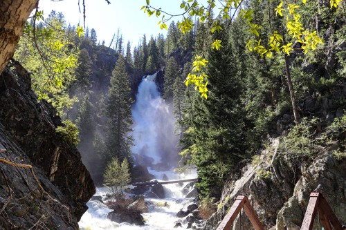 A waterfall cascades down rocky cliffs, surrounded by lush green trees and mist in a serene natural setting.