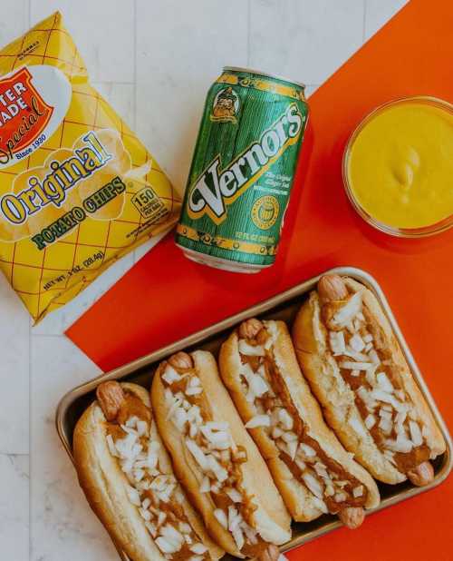 A tray of hot dogs topped with onions, a can of Vernors soda, a bowl of mustard, and a bag of potato chips.