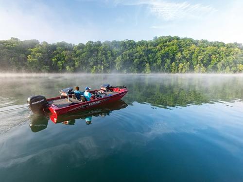 A red boat with two people glides across a misty lake, surrounded by lush green trees.