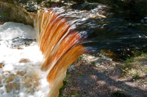 A cascading waterfall with orange-tinted water flowing over rocks, surrounded by greenery and dappled sunlight.