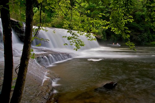 A serene waterfall cascades into a calm river, surrounded by lush green trees and a small boat in the distance.