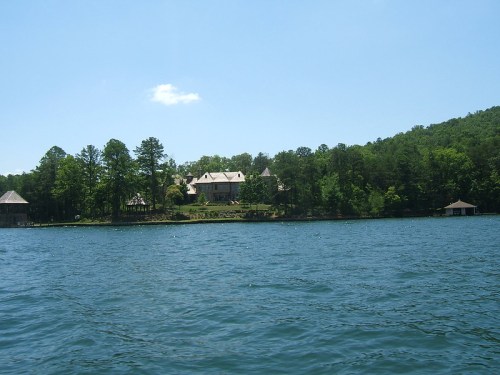 A serene lake view with a large house and lush green trees on the shore under a clear blue sky.