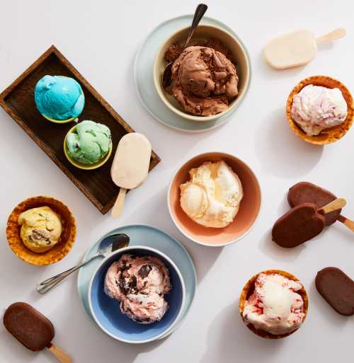 A variety of ice creams and popsicles in bowls and cones, featuring colorful flavors and textures on a white background.