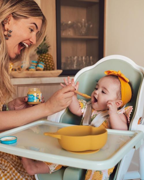 A joyful woman feeds a smiling baby in a high chair, both enjoying mealtime together. Bright, cheerful setting.