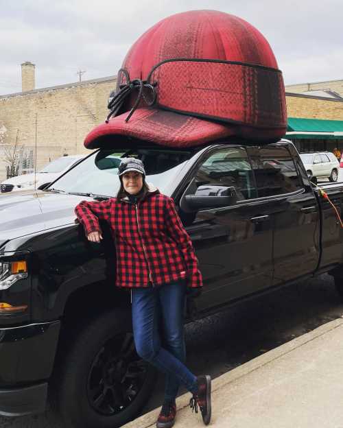 A person in a red plaid jacket leans against a truck topped with a giant red and black hat.