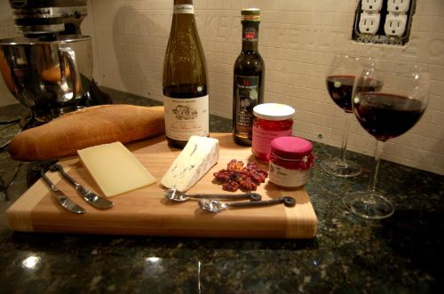 A wooden cheese board with various cheeses, wine, bread, and condiments, accompanied by two glasses of red wine.