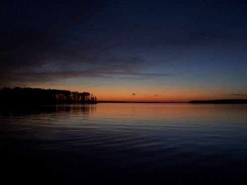 A tranquil lake at dusk, reflecting a gradient of orange and blue hues with silhouettes of trees along the shore.