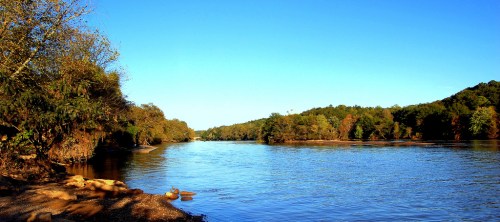 A serene river scene with clear blue skies, lush green trees, and calm water reflecting the landscape.