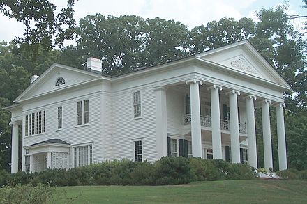 A large white historic house with tall columns and a manicured lawn, surrounded by trees under a cloudy sky.