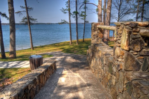 A stone pathway leads down to a serene lake surrounded by trees on a sunny day.