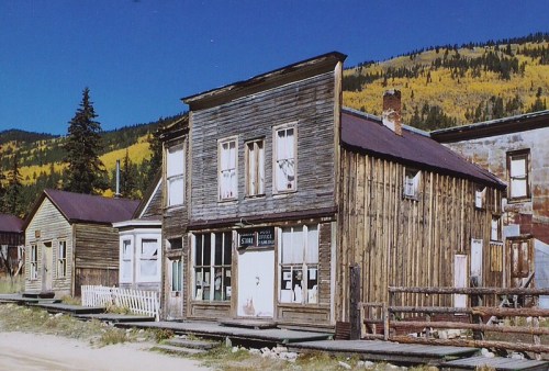 A weathered wooden building in a ghost town, surrounded by trees with autumn foliage under a clear blue sky.