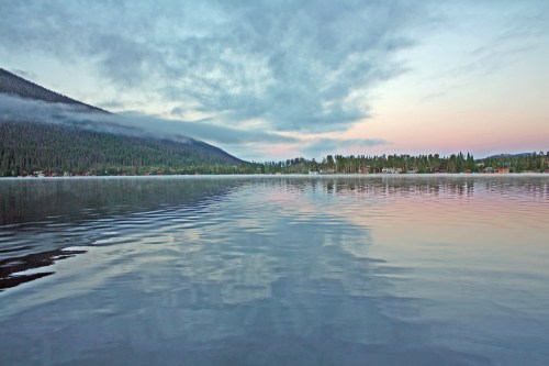 Serene lake scene at dusk, reflecting clouds and mountains, surrounded by lush trees and a calm atmosphere.