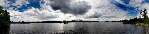 A panoramic view of a serene lake surrounded by trees and mountains under a cloudy sky.