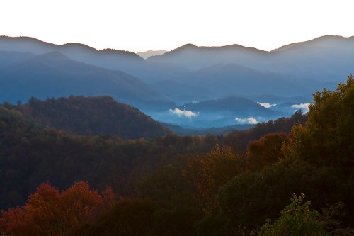 A serene mountain landscape at dawn, with layered peaks and mist rising from the valleys, showcasing autumn colors.