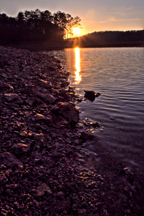 Sunset over a calm lake, with rocky shoreline and trees silhouetted against the colorful sky.
