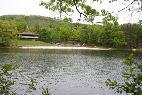 A serene lake surrounded by trees, with a sandy beach and a building in the background on a calm day.