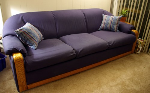 A purple sofa with wooden arms and two striped pillows, placed on a carpeted floor in a well-lit room.