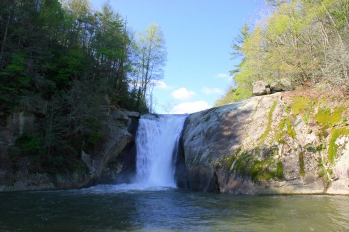 A serene waterfall cascades over rocky cliffs, surrounded by lush greenery and a clear blue sky.