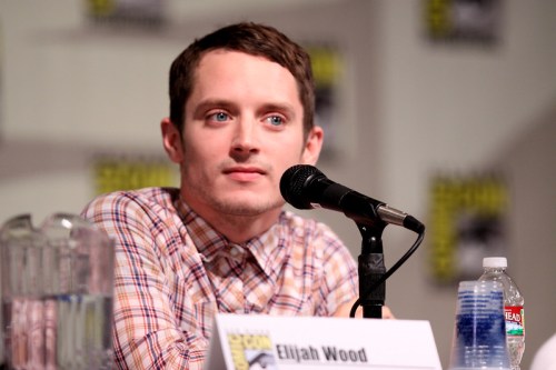 Elijah Wood at a panel discussion, speaking into a microphone with a water bottle and pitcher in the background.