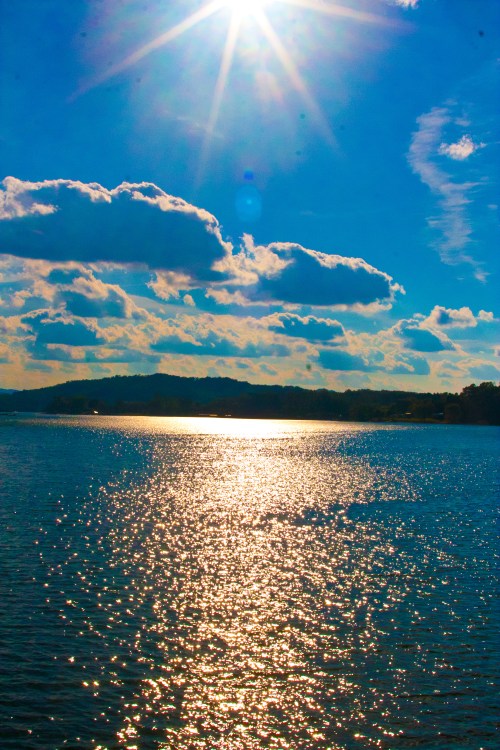 Bright sun reflecting on a shimmering blue lake, with fluffy clouds in a clear sky and distant hills on the horizon.