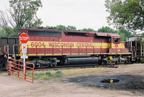 A Wisconsin Central locomotive parked near a stop sign and railroad crossing, surrounded by trees and tracks.