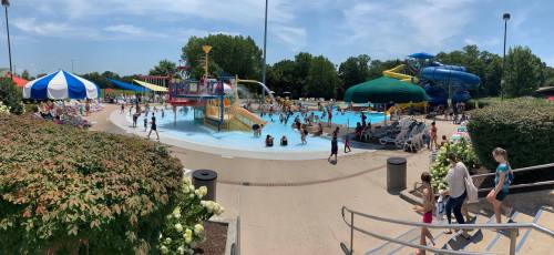 A busy water park with slides, a pool, and families enjoying the sunny day. Colorful tents and greenery surround the area.